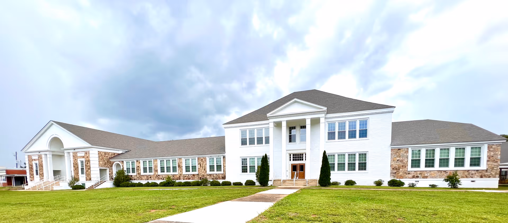 Front exterior of a large white two-story building with columns, many windows and a green lawn under a cloudy sky.