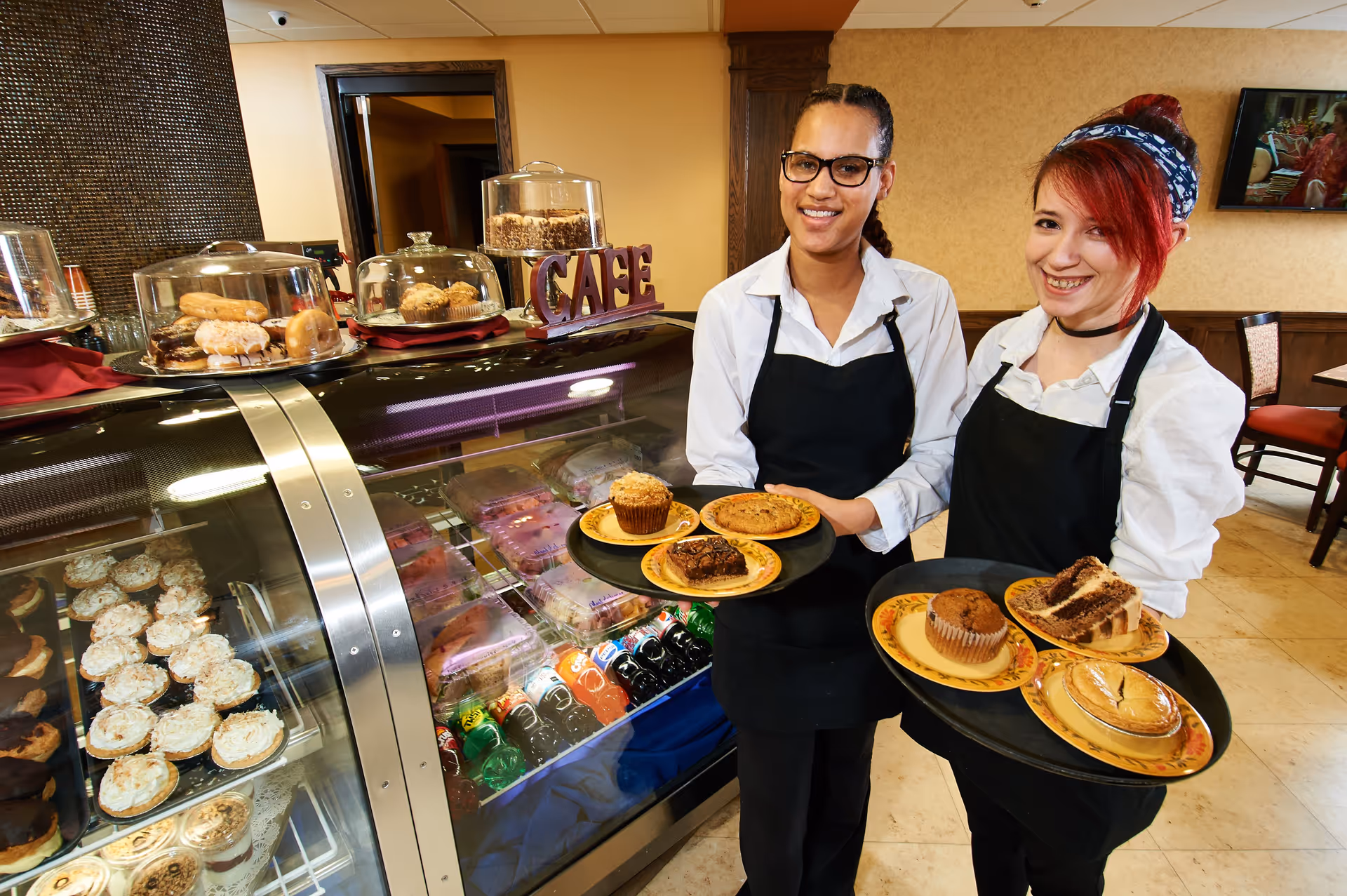 Two smiling cafe workers wearing black aprons and white shirts hold trays with various desserts including muffins, cookies, brownies, cake slices, and a pie in a cozy cafe setting with a display case of pastries and bottled drinks behind them.