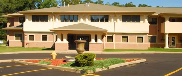 Two-story beige senior living facility with front entrance and center fountain in circular driveway.