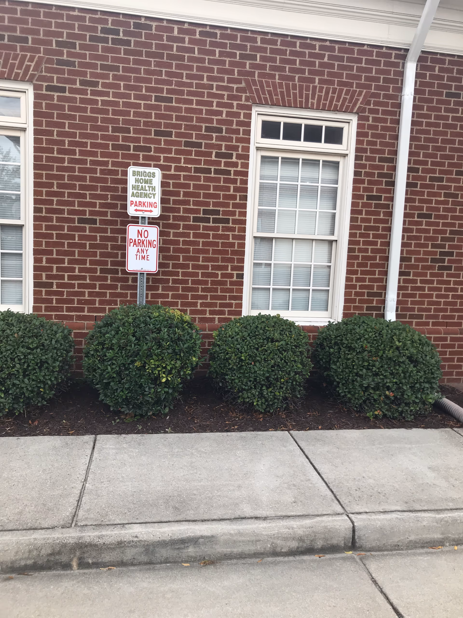 Brick building exterior with a window, trimmed shrubs, sidewalk, and parking signs.