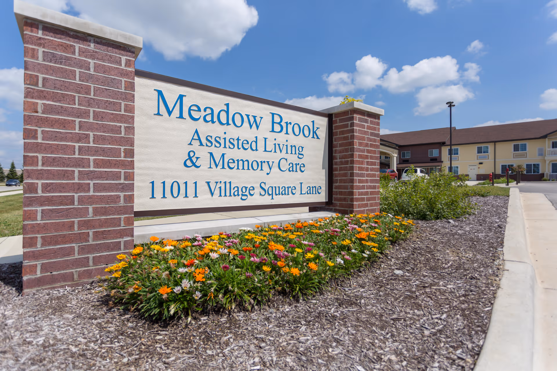 Outdoor view of a large sign for Meadow Brook Assisted Living & Memory Care at 11011 Village Square Lane, surrounded by a flower bed with colorful flowers and mulch, with the senior living facility building visible in the background under a partly cloudy blue sky.
