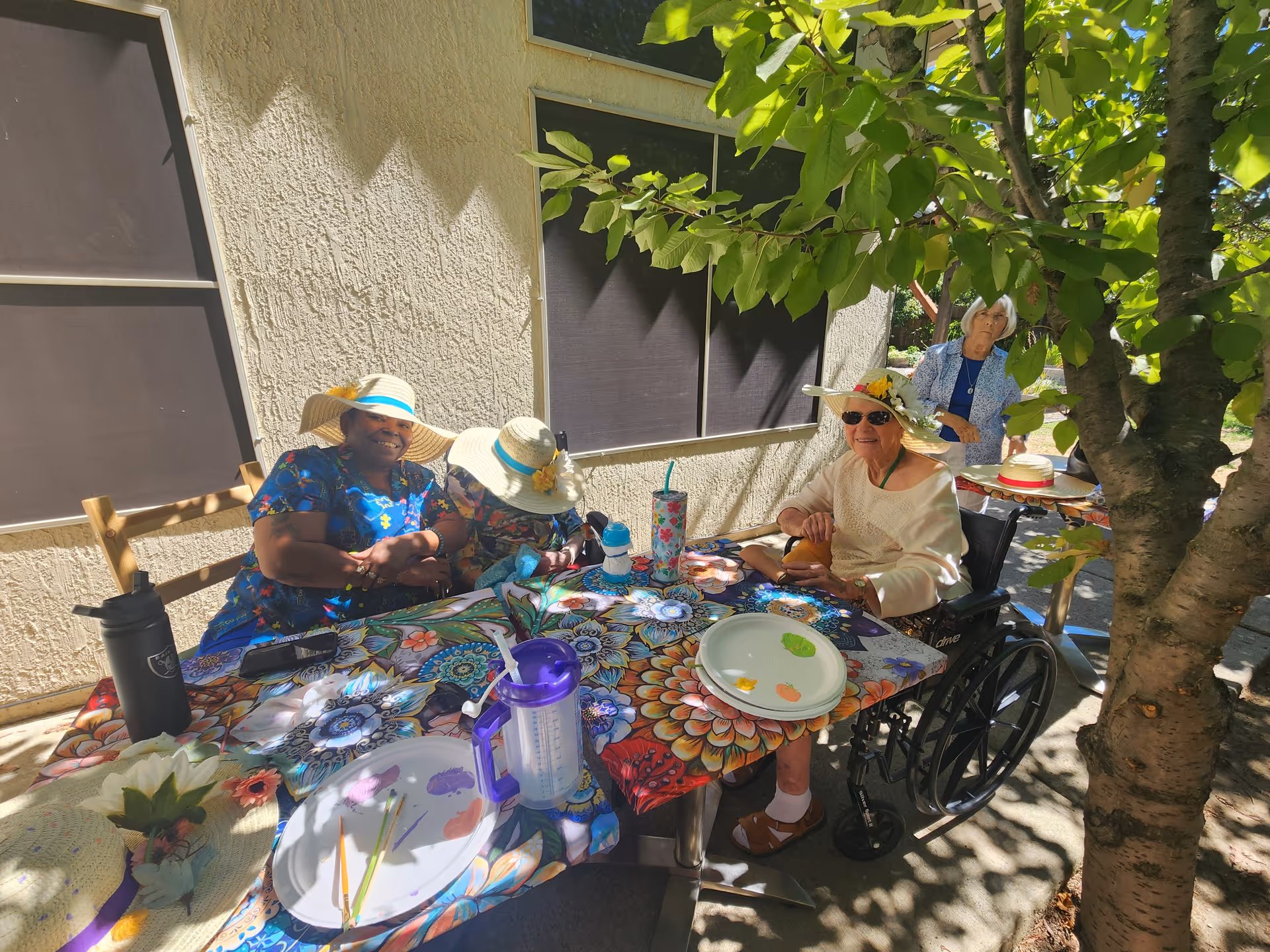 Senior residents wearing sun hats sit at a colorful outdoor table under a tree