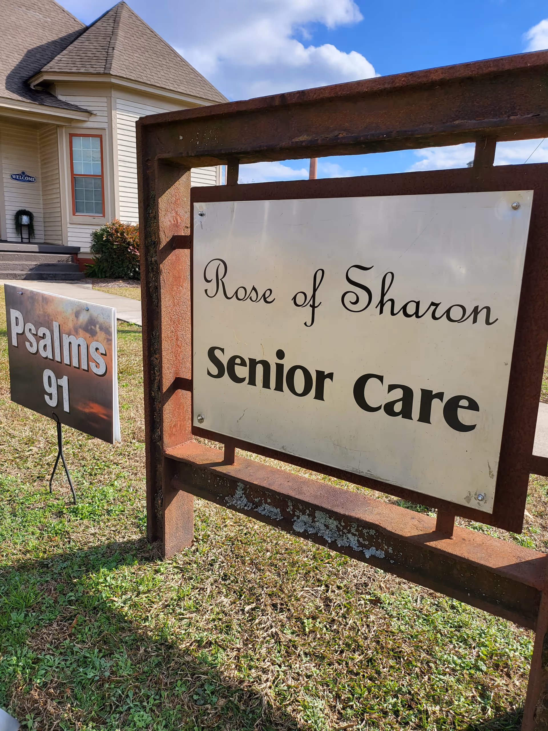 Outdoor sign reading "Rose of Sharon Senior Care" in front of a house with an additional "Psalms 91" sign on the lawn.