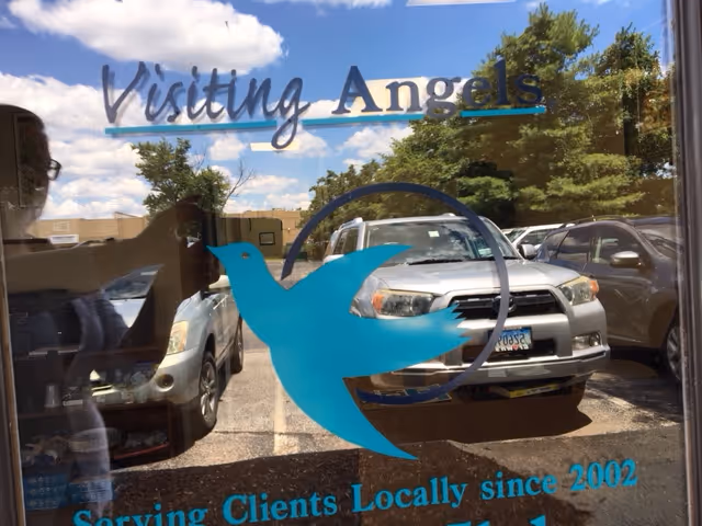 Glass storefront displays Visiting Angels sign with blue dove logo and reflections of parked cars.