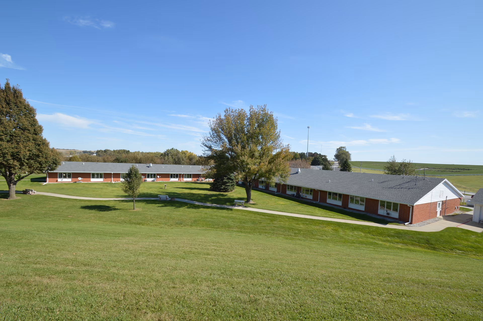 Exterior view of a single-story brick building with a gray roof surrounded by green grass and trees under a clear blue sky at Dunlap Specialty Care facility.