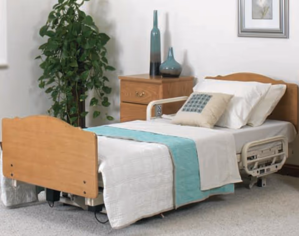Adjustable hospital-style bed in a clean bedroom with a wooden nightstand and plant.