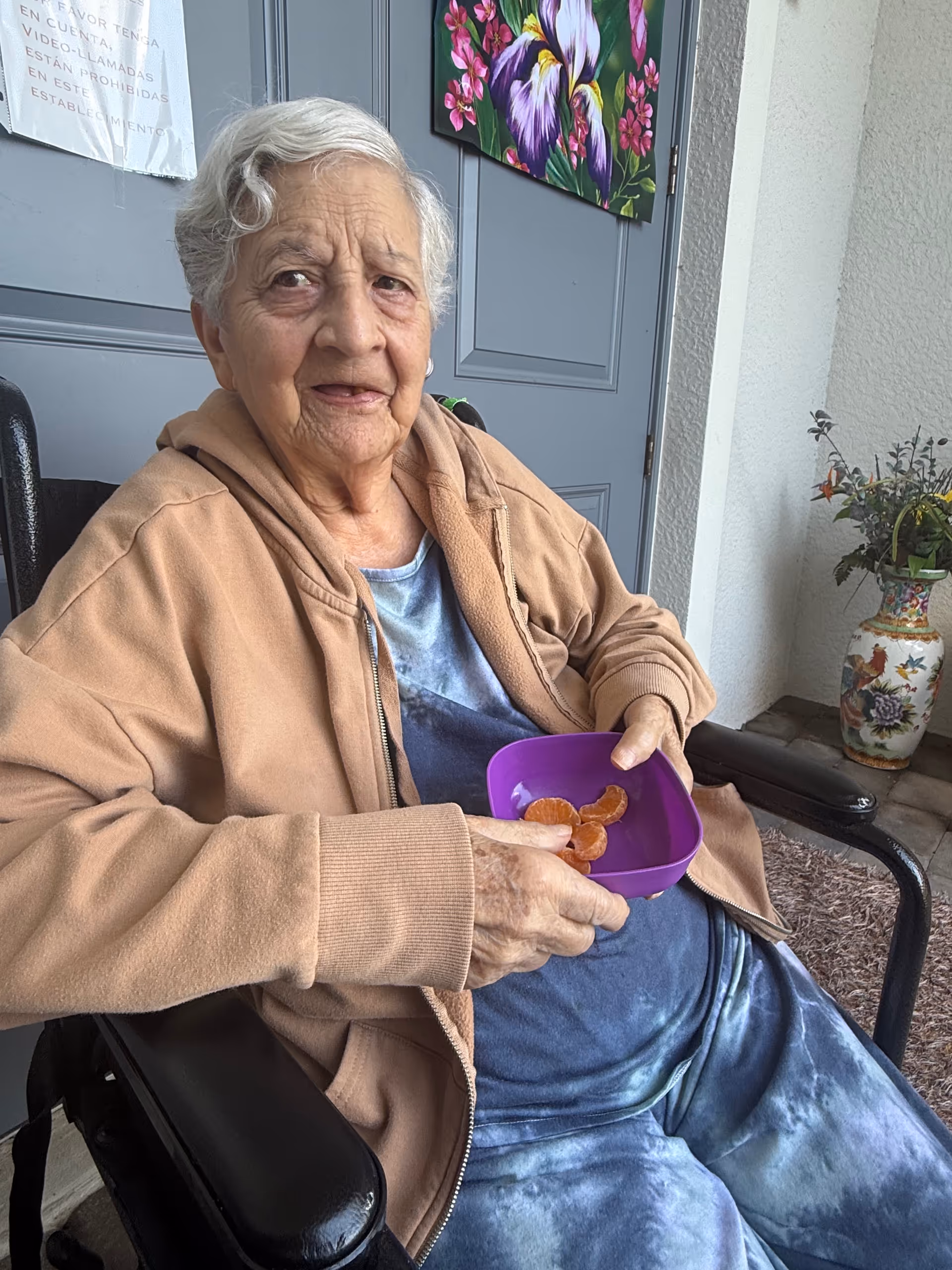 Elderly woman on a porch sits in a chair, holding a purple snack bowl.