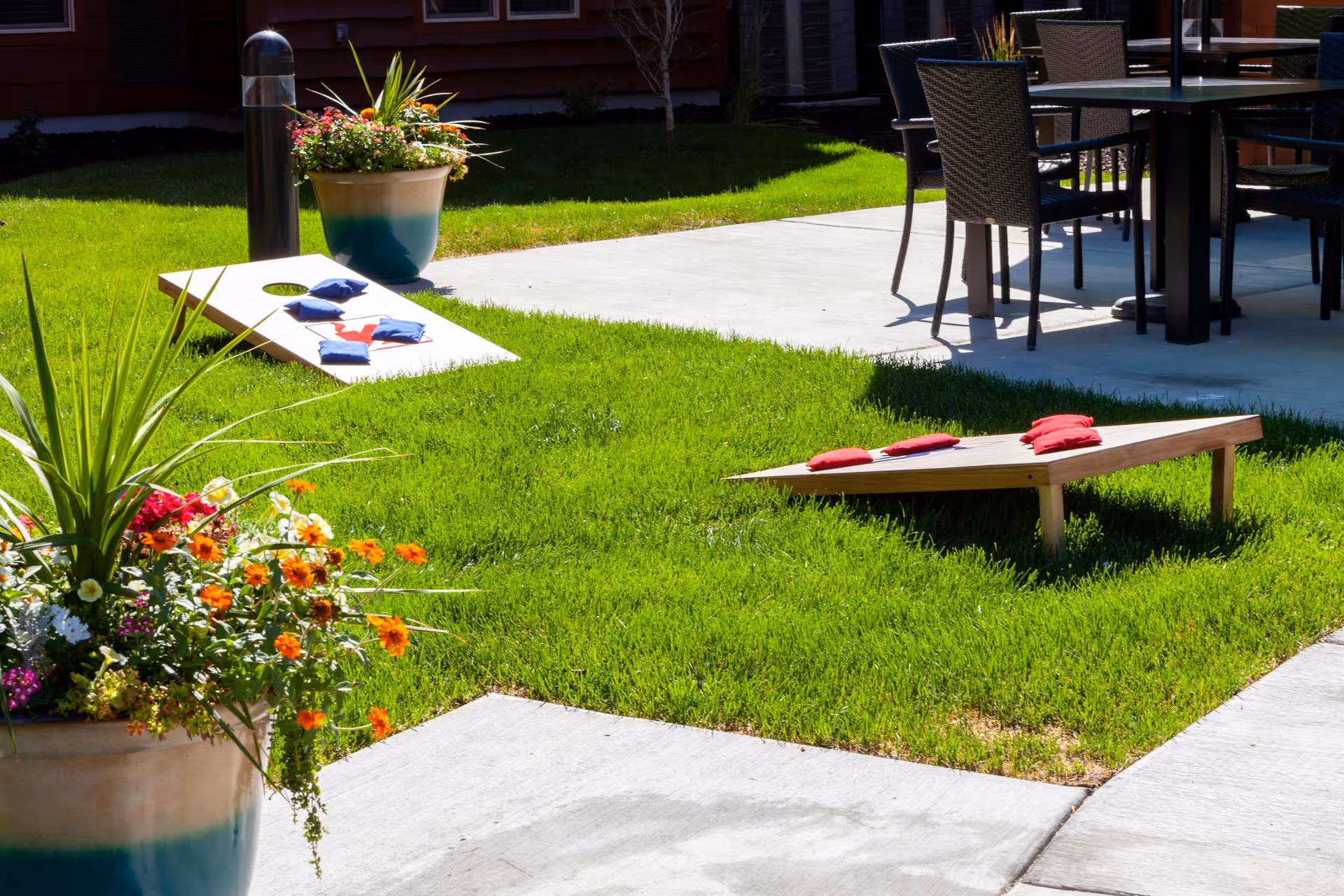 Outdoor patio area with green grass and two wooden cornhole boards set up for a game. There are colorful bean bags on the boards. Potted plants with flowers are placed near the grass, and a table with chairs is visible on a concrete patio in the background.