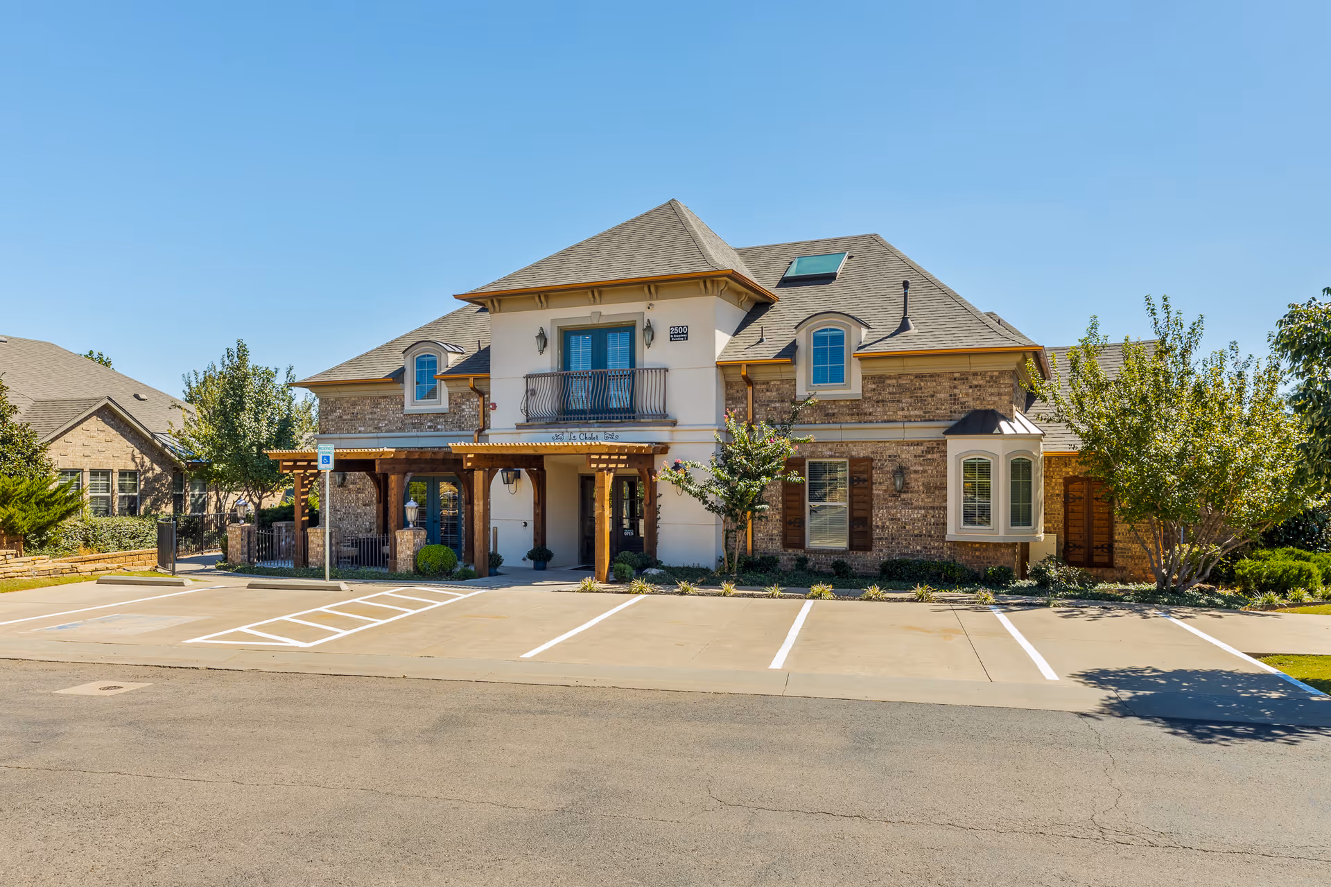 Exterior view of a two-story senior living facility building named Mon Abri, featuring a combination of brick and light-colored walls, a balcony, multiple windows, a covered entrance with wooden beams, and a parking area with marked spaces including a handicap spot. Trees and shrubs surround the building under a clear blue sky.