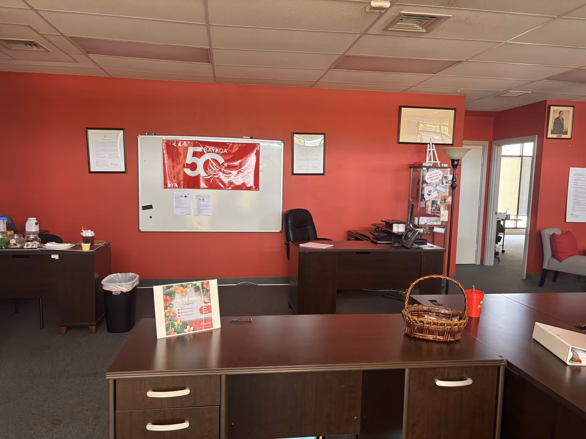Office reception area with red walls, desks, whiteboard, and framed documents.