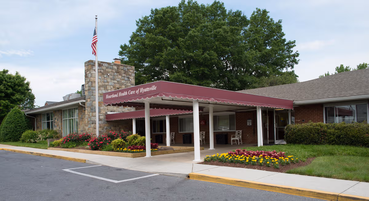 Senior living facility entrance with maroon canopy, stone column, and landscaped flower beds.