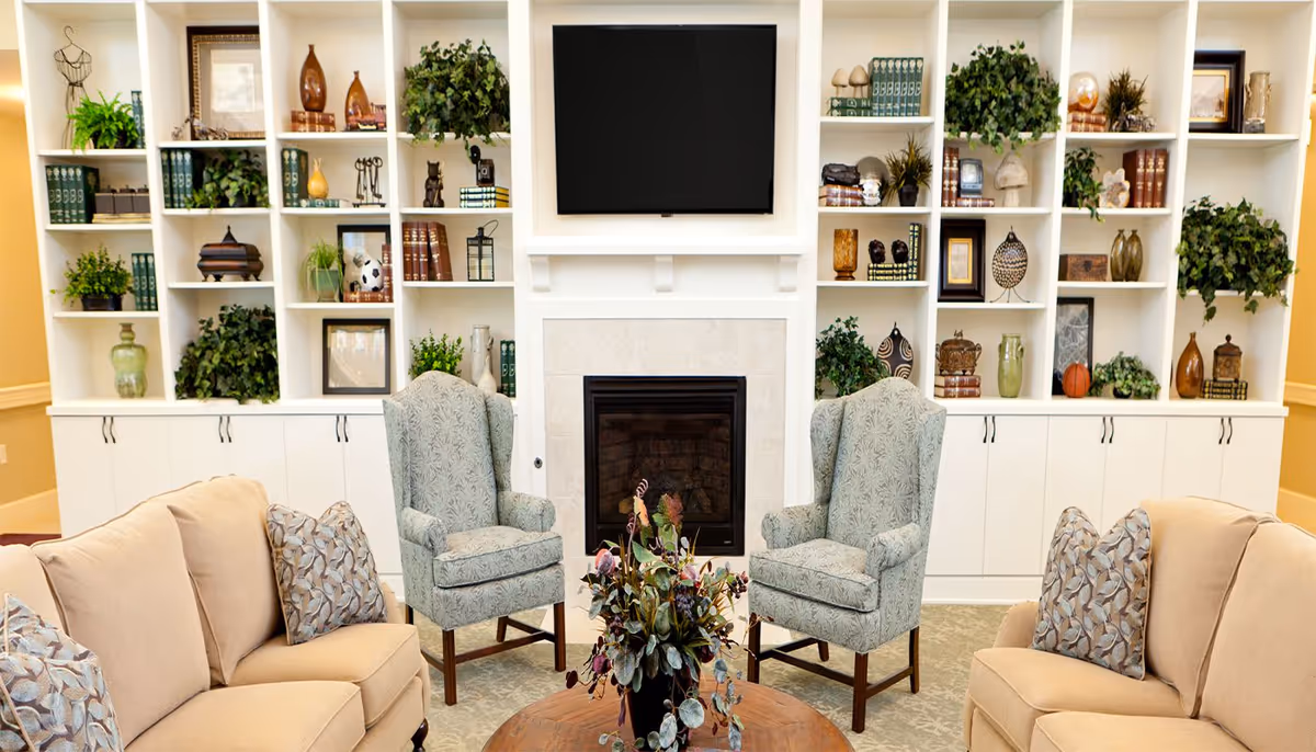 A cozy living room area with two beige sofas facing each other, each with patterned throw pillows, and two light blue patterned armchairs positioned near a fireplace. Above the fireplace is a mounted flat-screen TV. Surrounding the fireplace are white built-in shelves filled with various decorative items, books, and green plants.