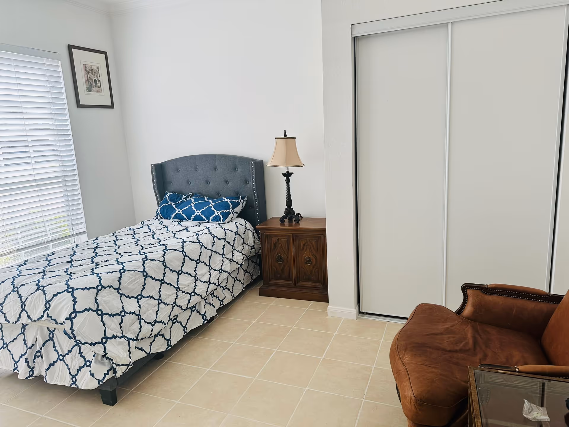 A single bedroom with a bed, nightstand, lamp, closet, and brown chair.