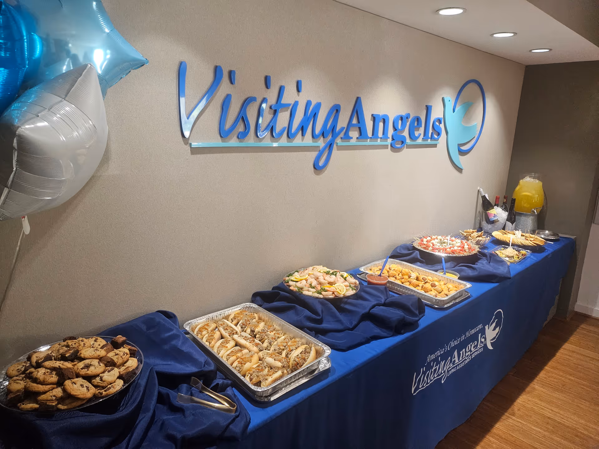Buffet table with pastries and snacks beside a blue Visiting Angels sign on the wall.