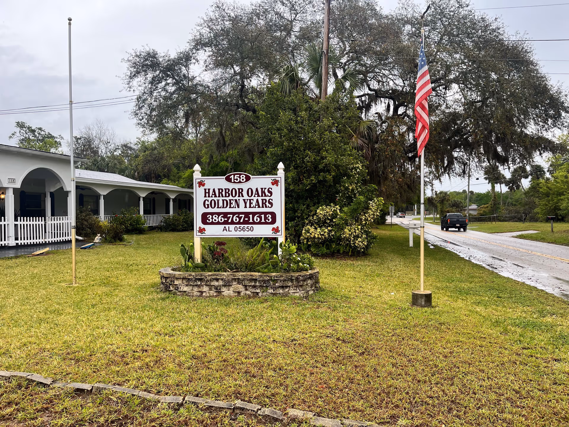 Exterior view of Harbor Oaks Golden Years facility with a white sign displaying the name, phone number, and license number. The building has a white picket fence and a covered porch with arches. There are two flagpoles in the grassy area, one with an American flag. Trees and a road with a vehicle are visible in the background under a cloudy sky.