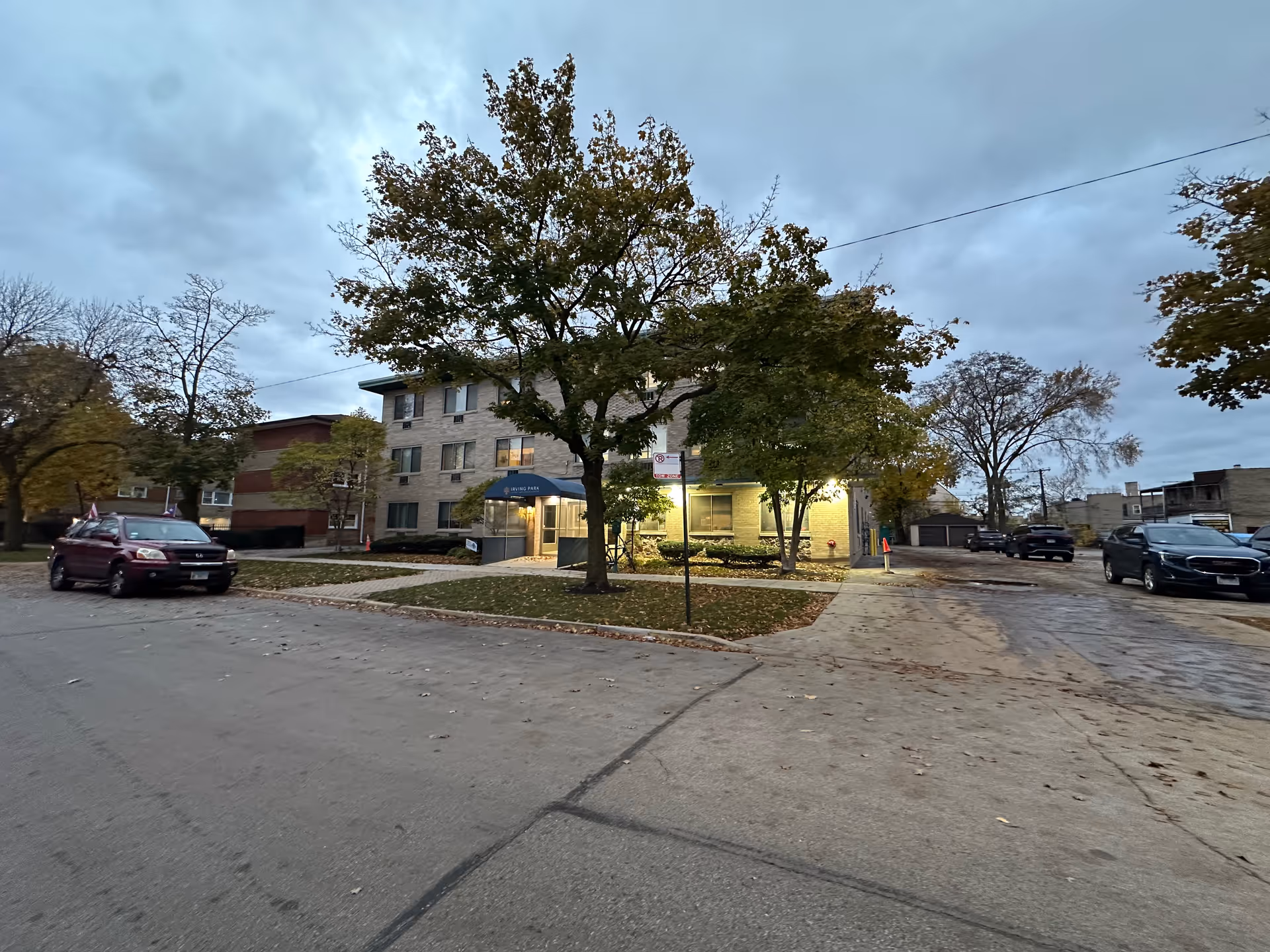 Exterior view of a three-story brick building with a blue awning over the entrance labeled Irving Park. Several trees with autumn foliage are in front of the building, and cars are parked along the street and driveway. The sky is cloudy and it appears to be early evening.
