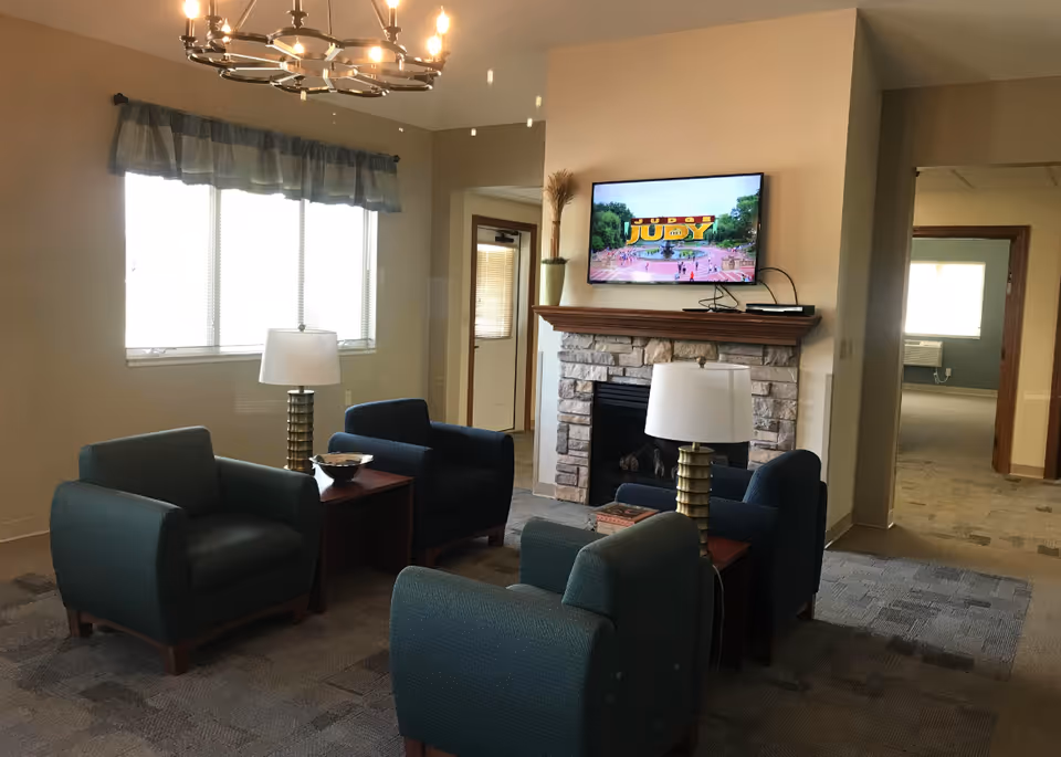A cozy senior living lounge with blue chairs, a stone fireplace, and a mounted TV.