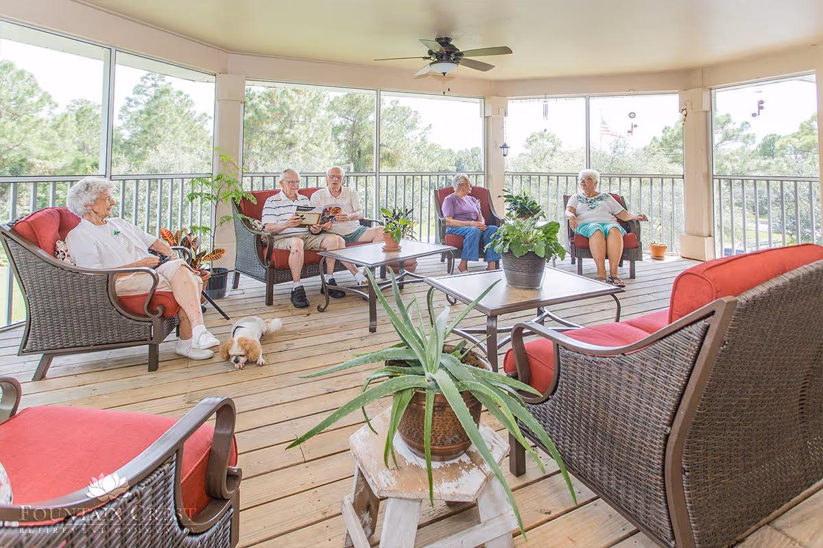 A group of elderly people sitting on cushioned wicker chairs on a covered wooden porch with potted plants and a small dog lying on the floor. The porch has a ceiling fan and overlooks a green outdoor area with trees.