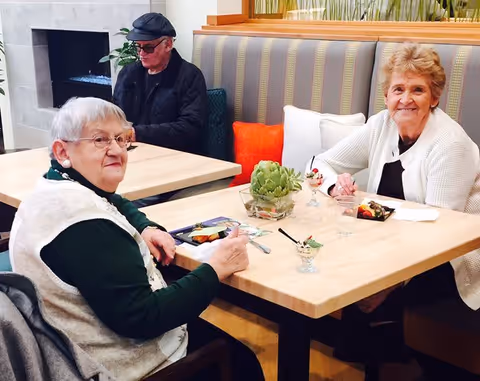 Two elderly women sitting at a wooden table in a dining area, enjoying dessert. One woman is wearing a white sweater and the other a white vest over a green shirt. In the background, an elderly man wearing a black cap and jacket is seated at another table near a modern fireplace. The seating area has striped upholstery and colorful cushions.