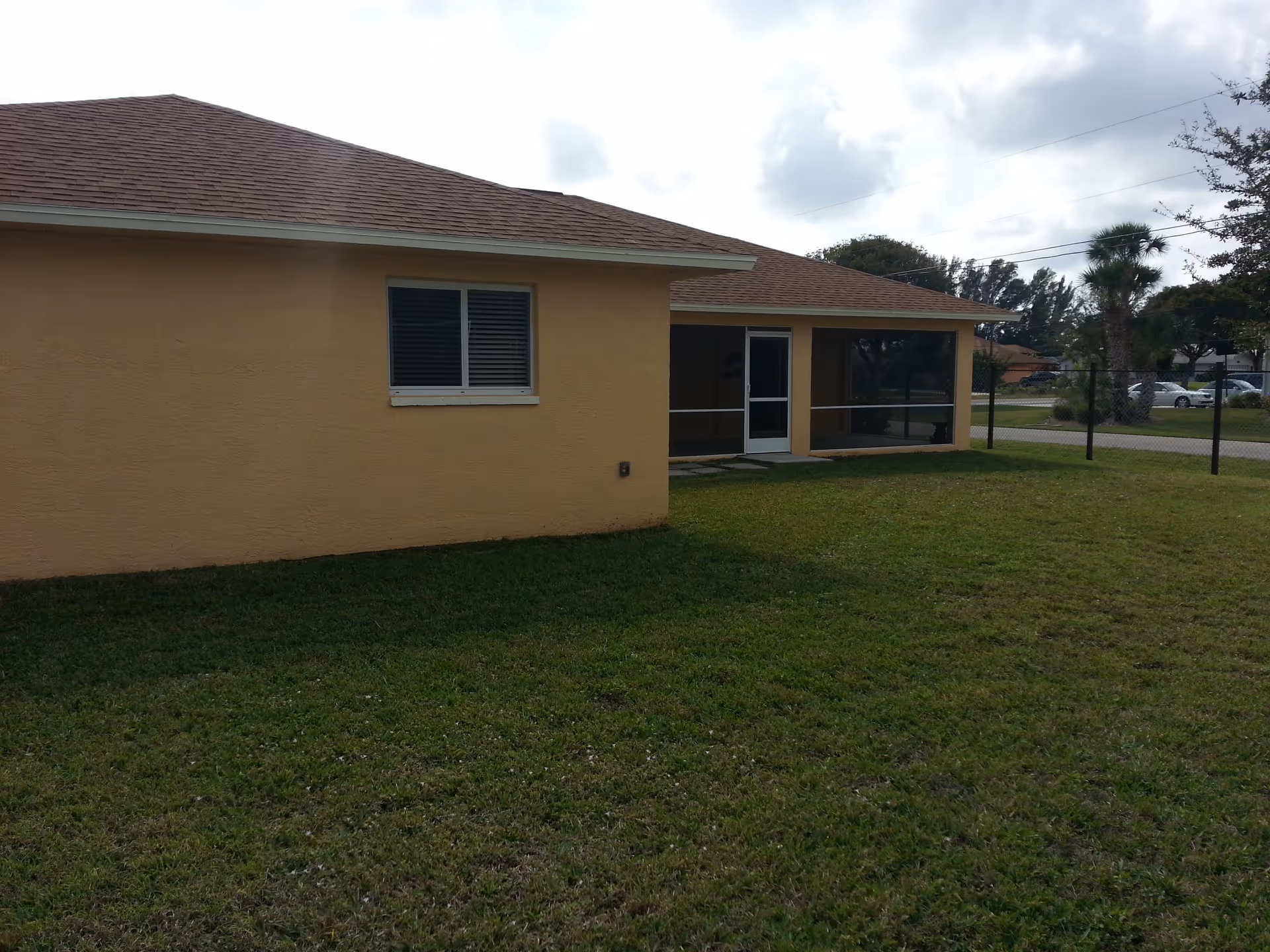 Exterior view of a single-story building with beige walls and a brown shingled roof. There is a window with closed blinds on the left side and a screened porch with a door on the right side. The building is surrounded by a grassy yard and a chain-link fence. Trees and parked cars are visible in the background under a cloudy sky.