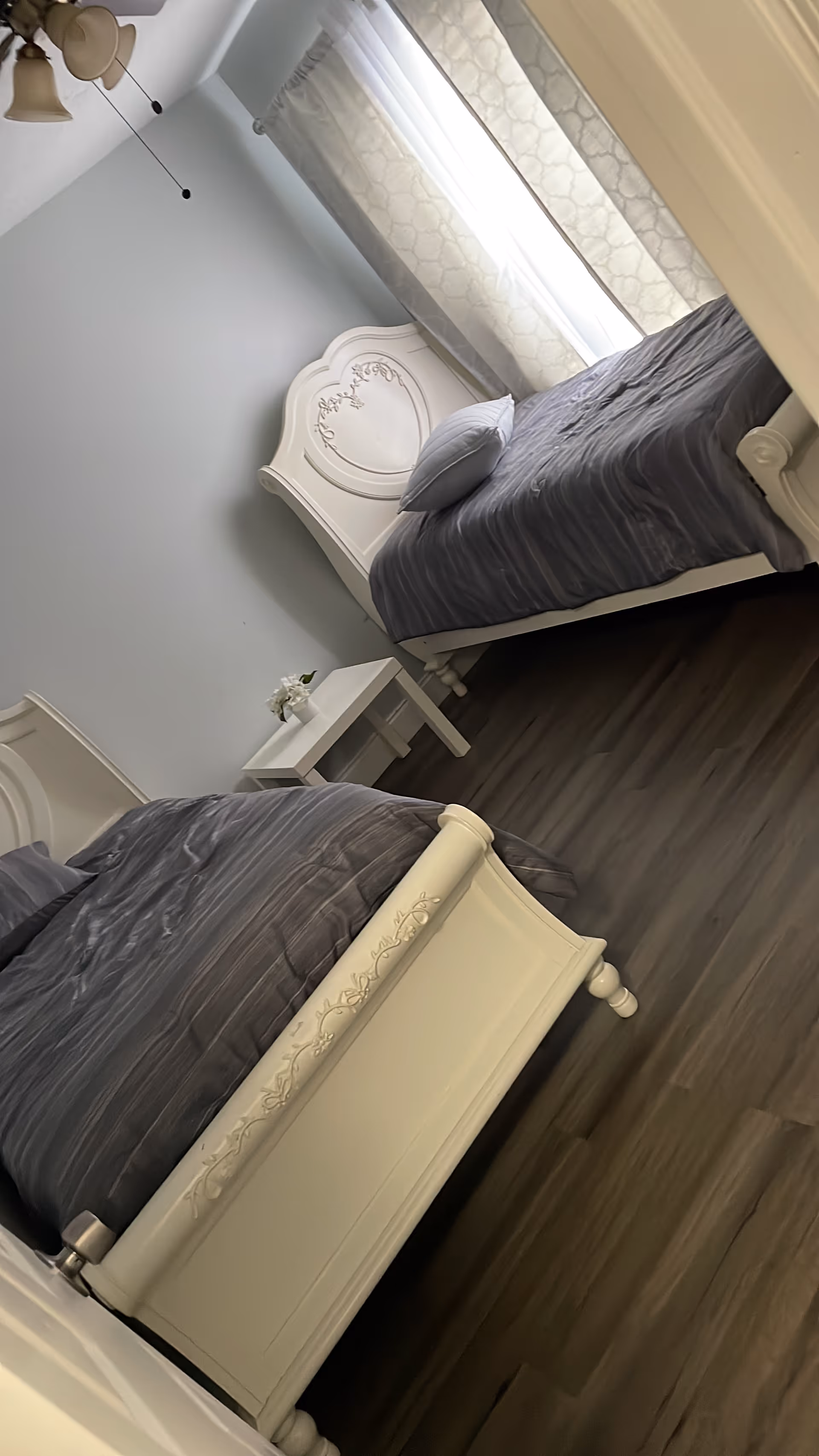 Two white ornate twin beds flank a small nightstand under gray curtains.