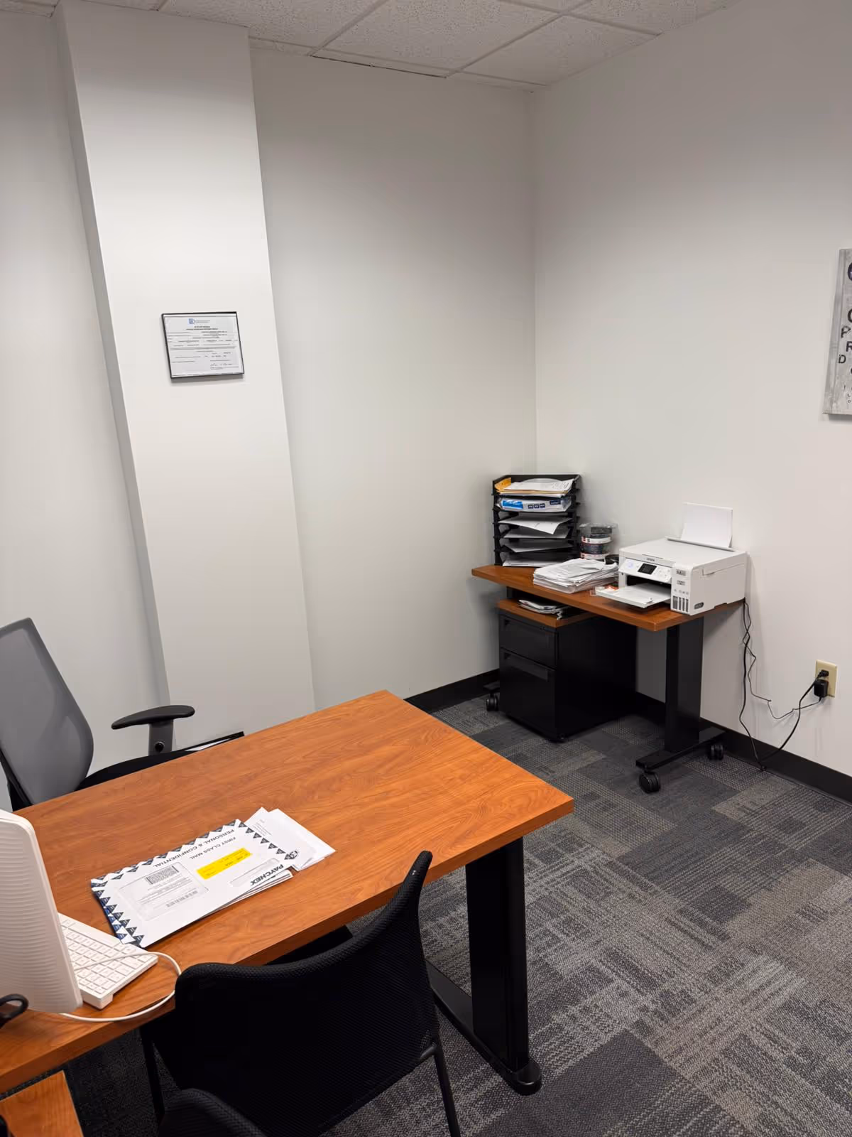 Office corner with wooden desk, computer, printer, and filing tray in a small room.