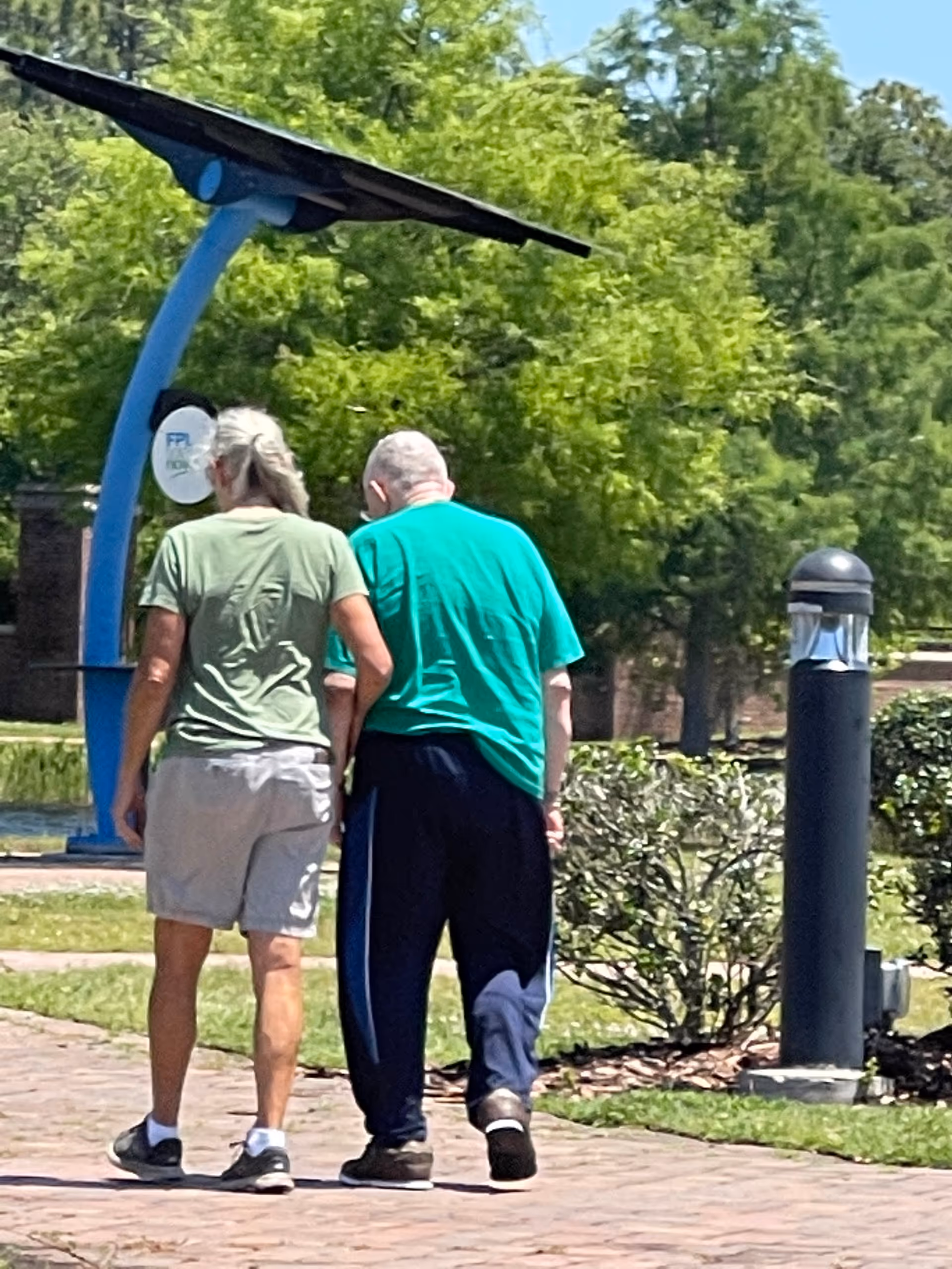 Two elderly residents walk along a sunny brick path in a landscaped outdoor area.