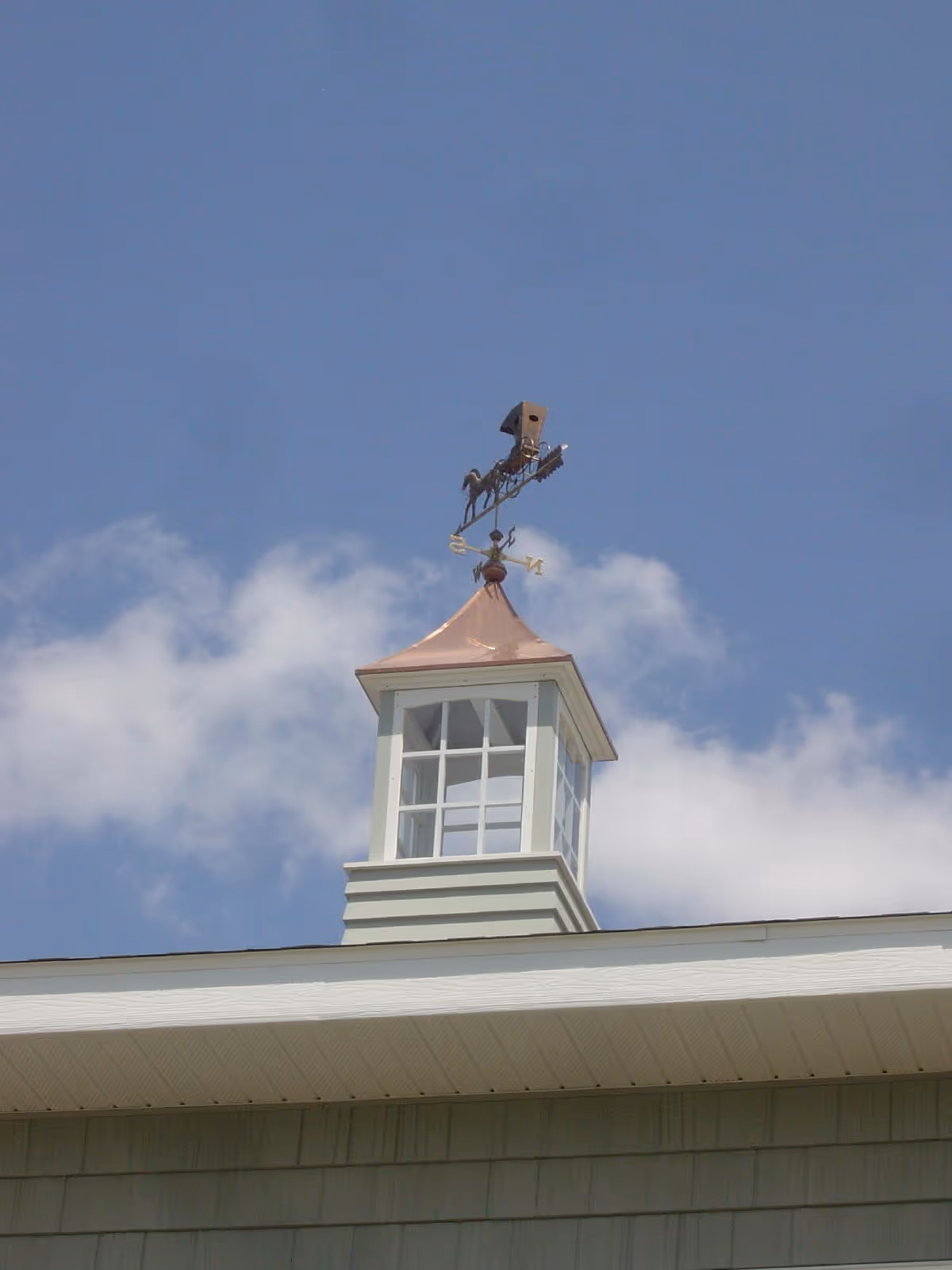 Small cupola with windows and a copper roof topped by a decorative weather vane against a blue sky.