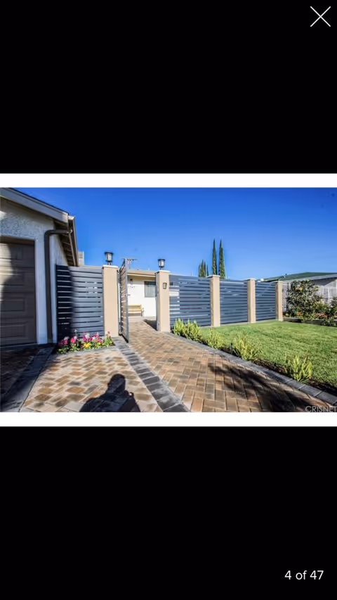 Paved walkway leading to a gated entrance of a residence with a modern horizontal slat fence, green lawn, and some plants under a clear blue sky.