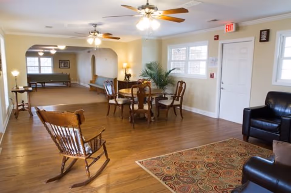 Bright communal living room with a wooden rocking chair, round dining table and chairs, sofas, ceiling fans, and an exit door.