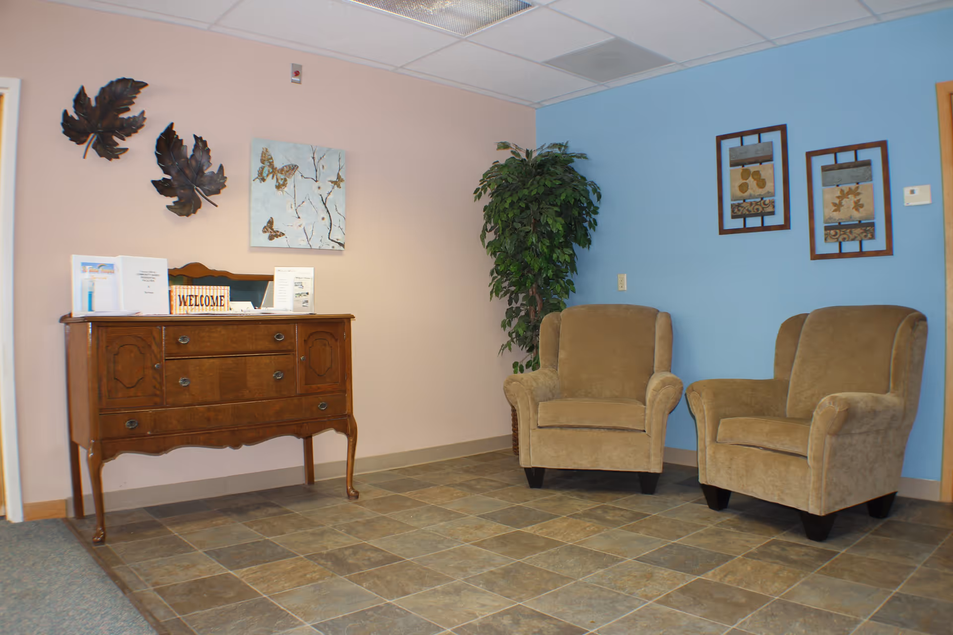 A cozy waiting area with two beige upholstered armchairs placed against a blue wall. To the left, there is a wooden sideboard with a mirror, decorative items including a 'WELCOME' sign, and some papers. The walls are painted in light pink and blue, adorned with framed artwork and metal leaf decorations. A potted plant is positioned between the chairs and the sideboard. The floor is tiled with a mix of brown and gray tones.
