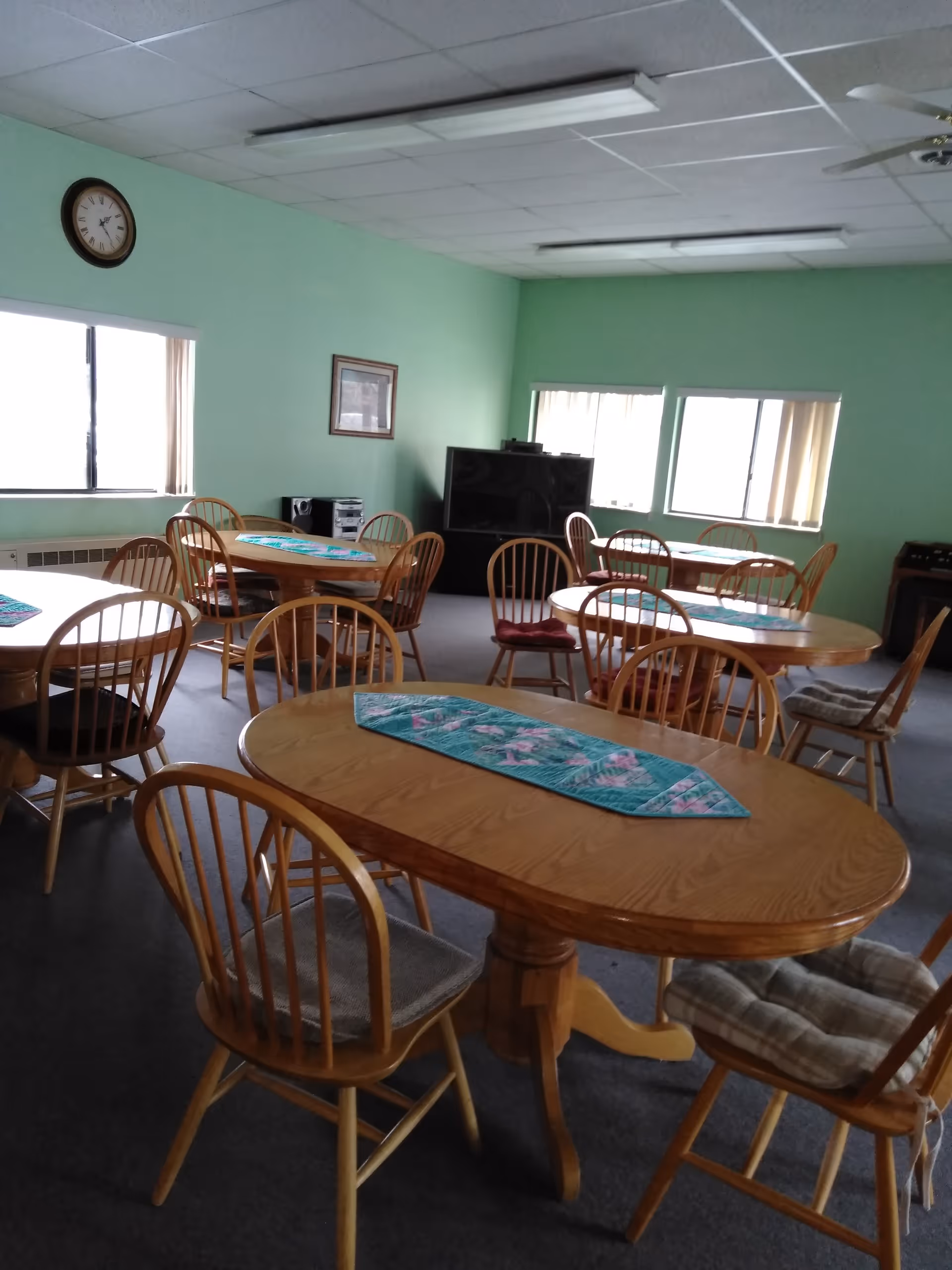 A common room with multiple wooden tables and chairs arranged around them. Each table has a decorative runner. The walls are painted light green, and there are several windows with vertical blinds letting in natural light. A clock is mounted on one wall, and a large television sits on a stand in the corner of the room.