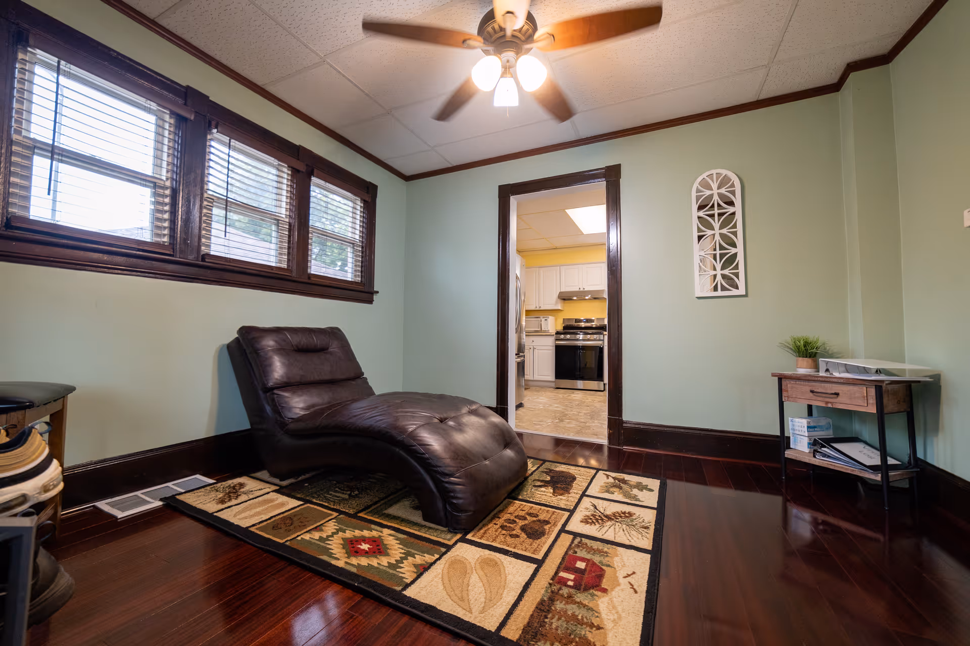 Cozy living area with a leather recliner, patterned rug, ceiling fan, and kitchen doorway.