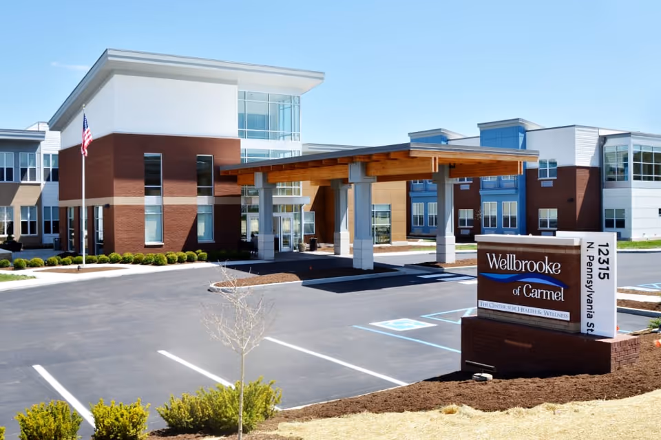 Exterior view of Wellbrooke of Carmel, a senior living facility, showing a modern two-story building with large windows, a covered entrance with wooden beams, an American flag, and a parking lot with marked spaces including handicapped spots. A sign in front displays the facility name and address.