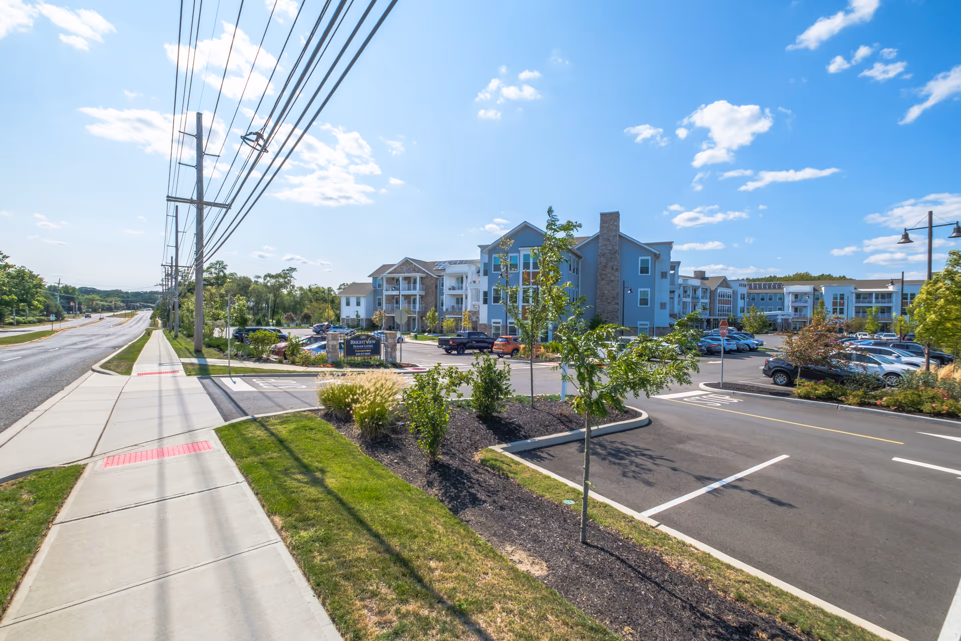 Street-level view of a senior living complex with a sidewalk, parking lot, landscaped islands, and multi-story buildings under a blue sky.