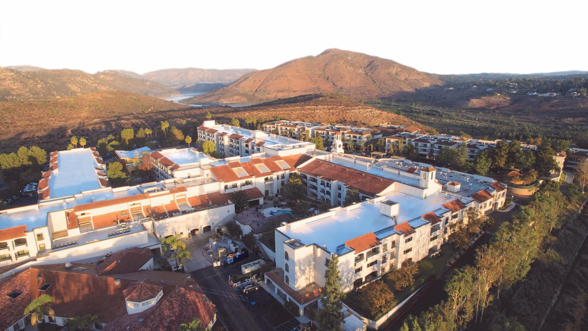 Aerial view of Casa de las Campanas facility surrounded by hills and trees, showing multiple connected buildings with white walls and red-tiled roofs under a clear sky.