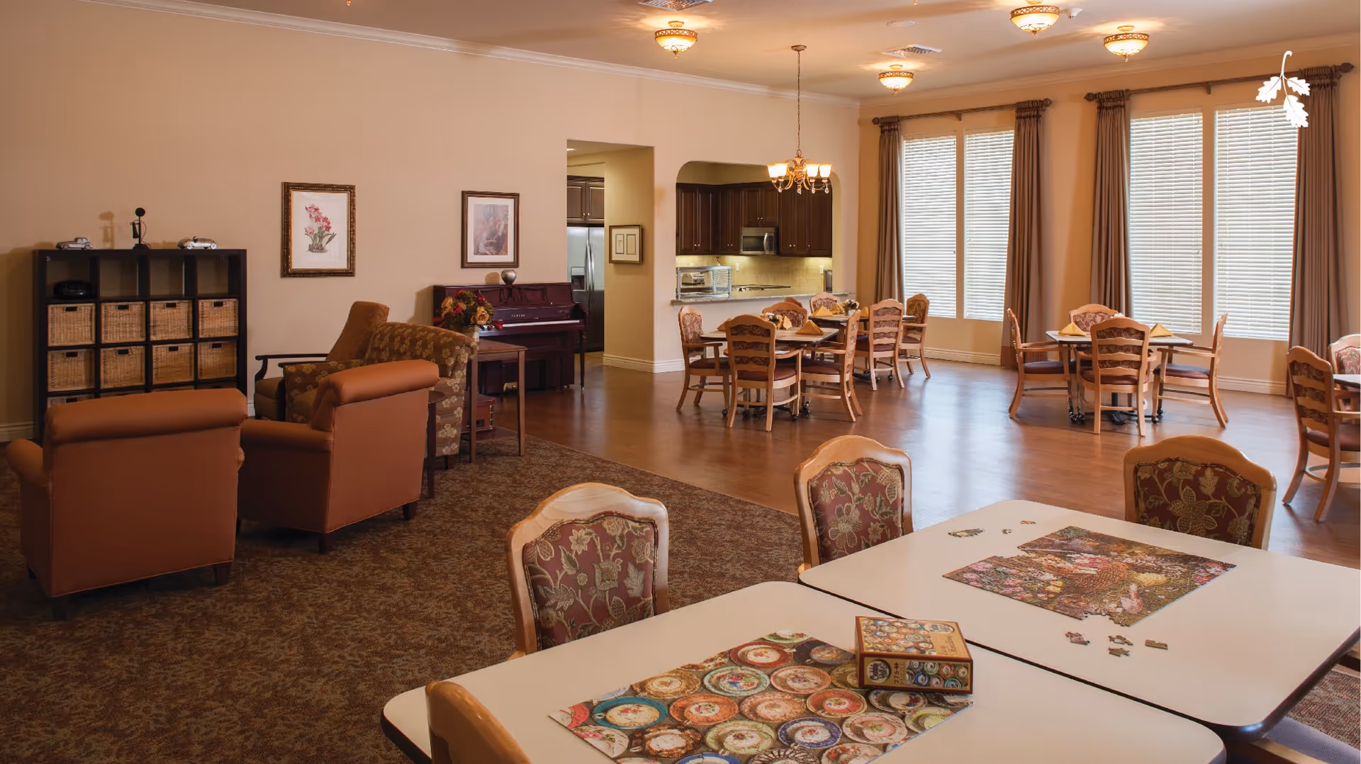 Spacious memory care common area with dining tables, comfortable seating, and a visible kitchen in back.