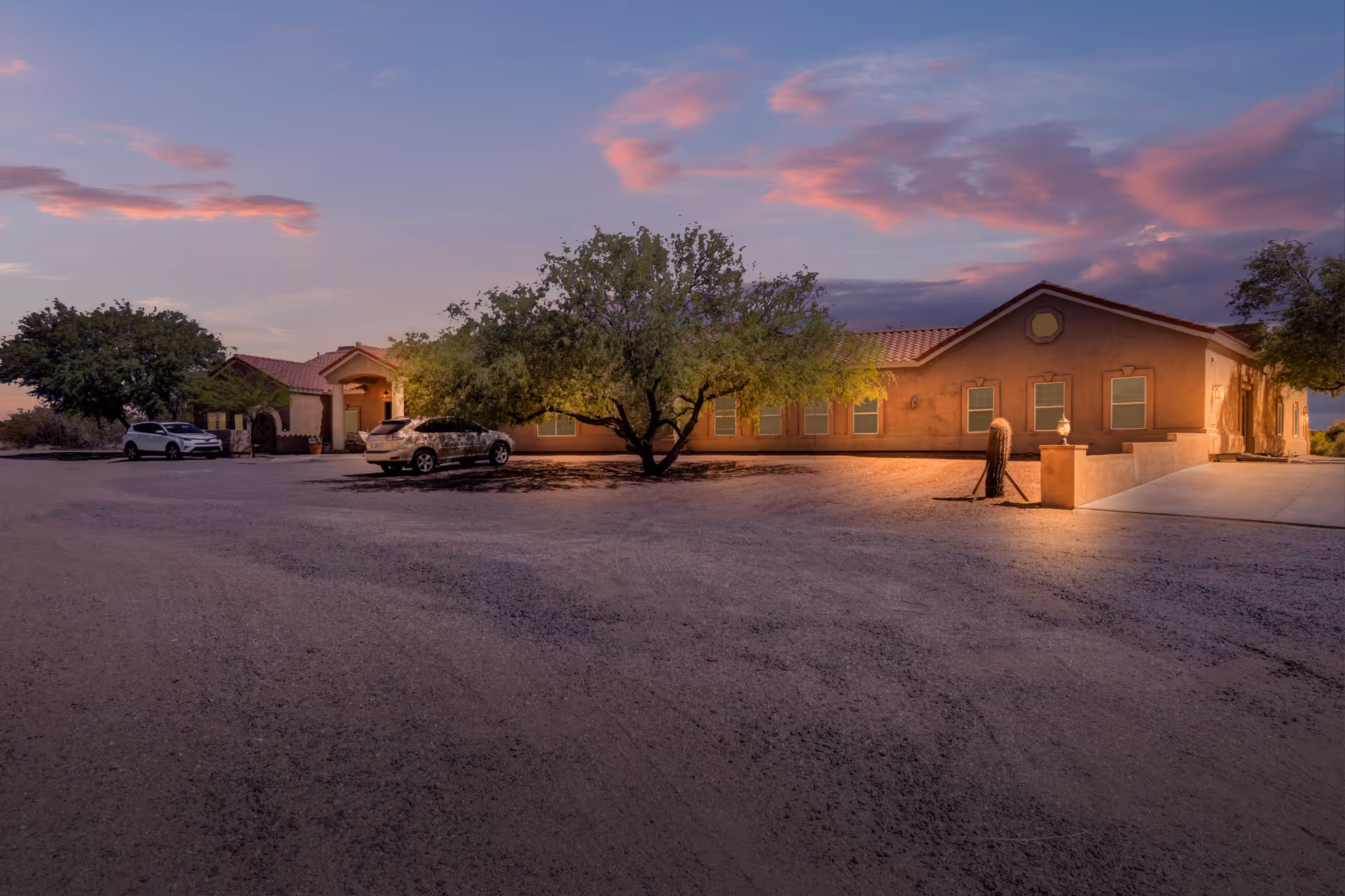 Exterior view of Lovin Manor facility at dusk with a gravel parking area, a few parked cars, and a large tree in front of a single-story building with a tiled roof and multiple windows. The sky is partly cloudy with pink and purple hues from the setting sun.