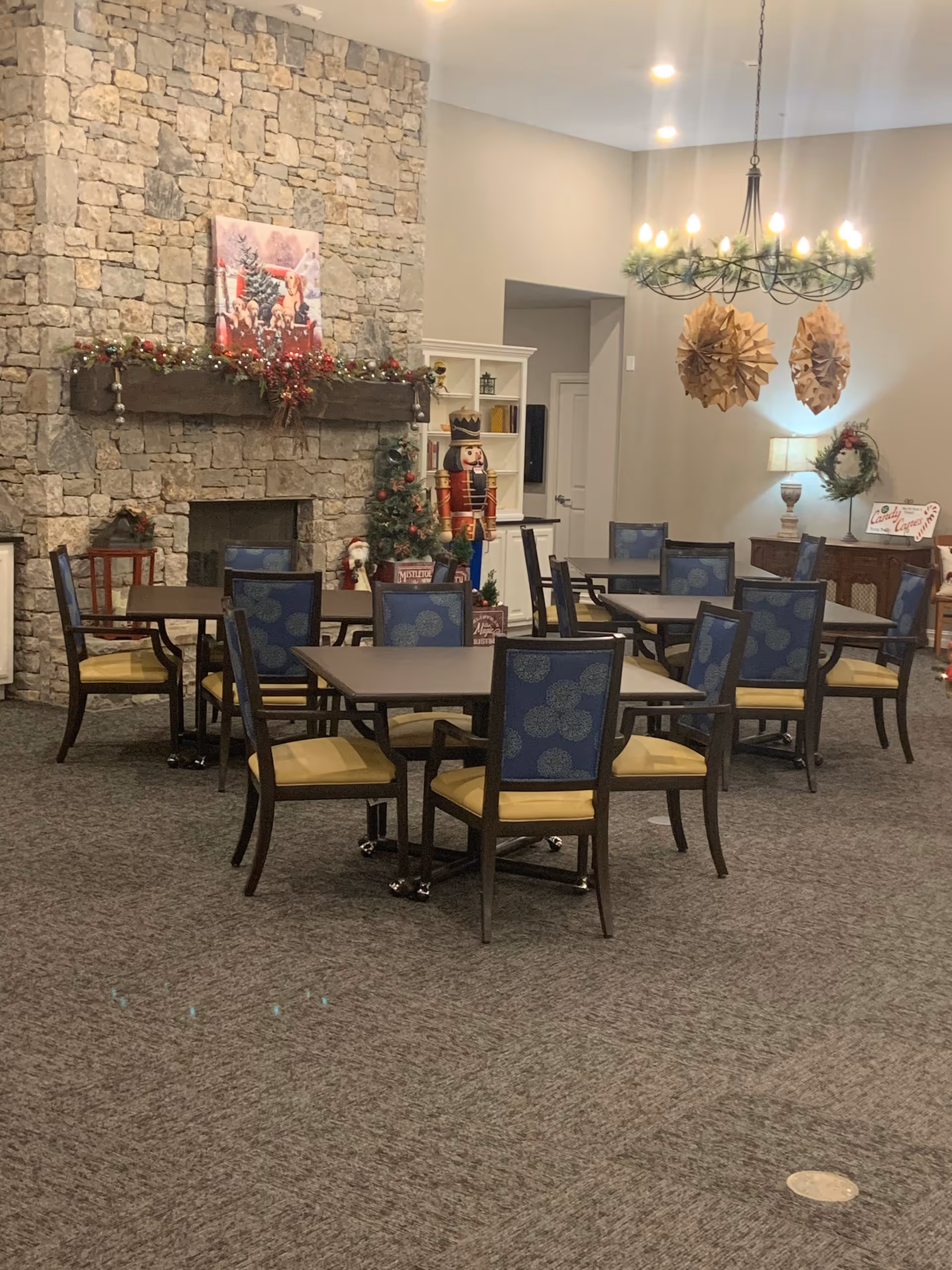 A cozy dining area with multiple tables and chairs arranged on a carpeted floor. The room features a large stone fireplace decorated with Christmas garlands and ornaments. There is a Christmas tree and a large nutcracker figure near the fireplace. A chandelier with warm lights hangs from the ceiling, and festive decorations are visible throughout the room.