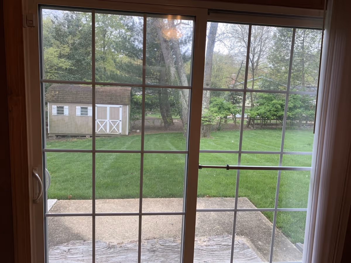 View through a sliding glass door with white grid frames, looking out onto a green lawn with trees and a beige shed with white trim in the background.