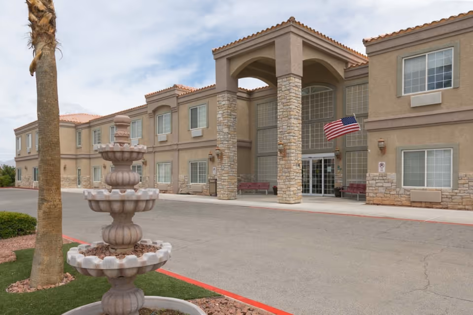 Exterior view of Desert View Senior Living facility showing a two-story building with beige walls, stone accents, and a tiled roof. There is a large entrance with tall stone pillars and an American flag mounted near the door. In the foreground, there is a tiered stone fountain and a palm tree on a landscaped area with grass and rocks.