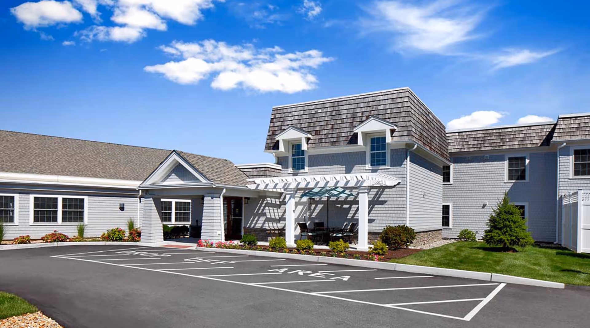 Front exterior of a white two-story senior living building with a covered entrance, pergola seating area, landscaping, and marked parking spaces under a blue sky.