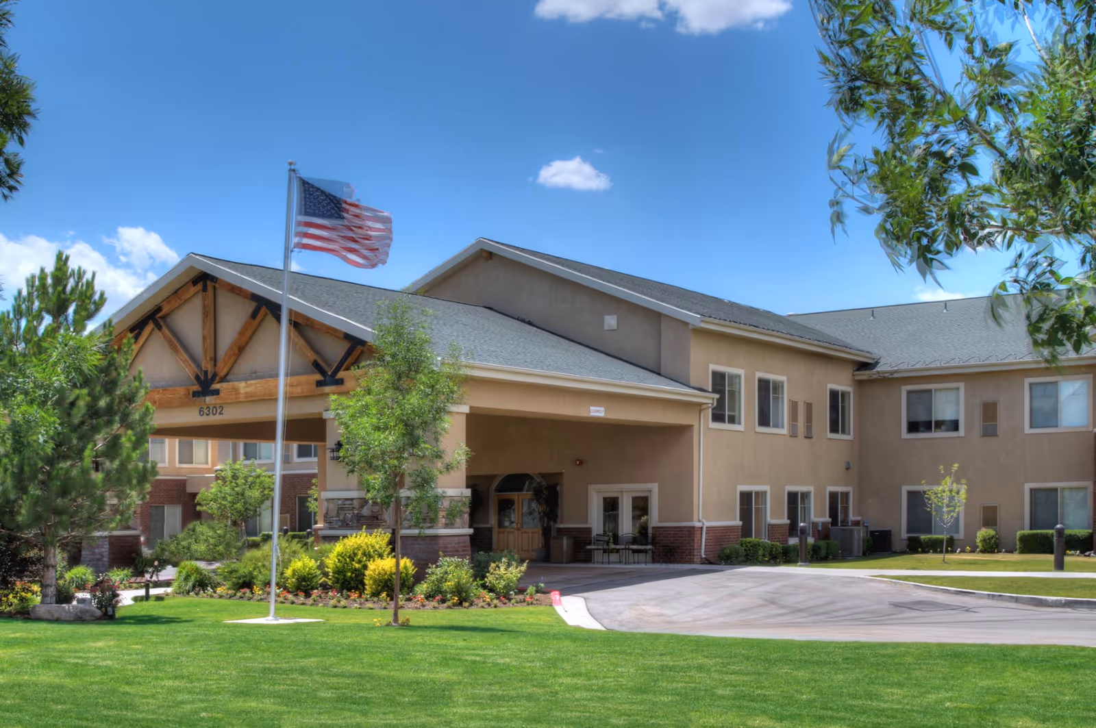 Exterior view of a senior living facility building with beige walls and a gray roof under a clear blue sky. There is a covered entrance supported by stone and brick pillars, an American flag on a flagpole in front, and well-maintained green grass and landscaping around the building.