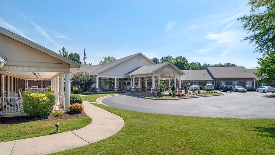 Exterior view of Brookdale New Hope senior living facility showing a single-story building with a covered entrance, a circular driveway, several parked cars, a well-maintained lawn, and an American flag on a flagpole under a clear blue sky.