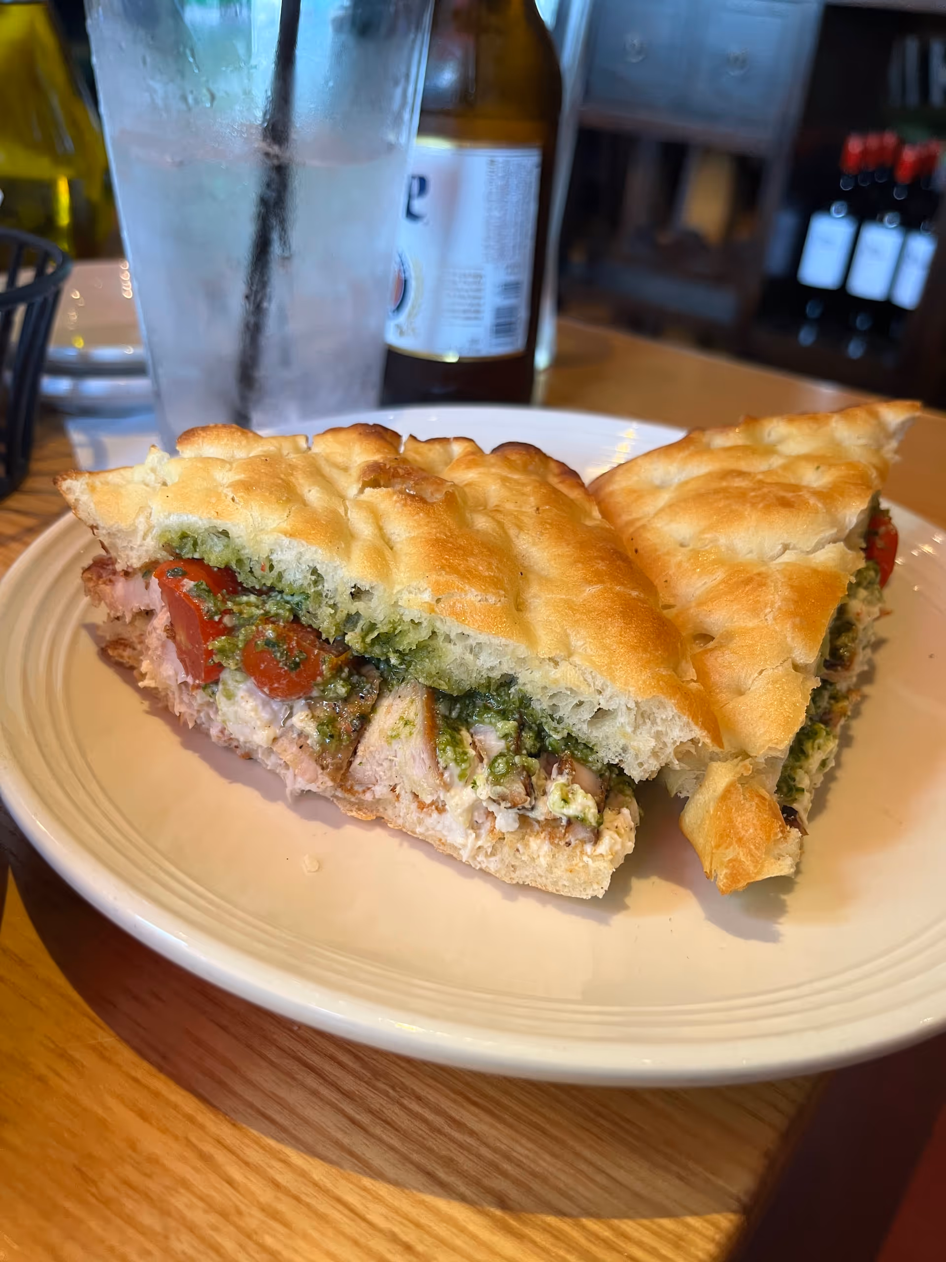 A sandwich on a plate with a bottle and glass at a dining table.