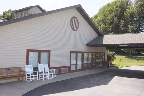Front entrance of a single-story white building with rocking chairs and a covered drop-off driveway.