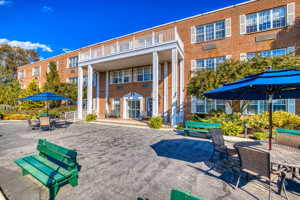 Exterior view of Amber Court senior living facility showing a large brick building with white columns at the entrance. There are green benches and outdoor tables with blue umbrellas on a paved area in front of the building. Trees and shrubs surround the area under a bright blue sky with some clouds.