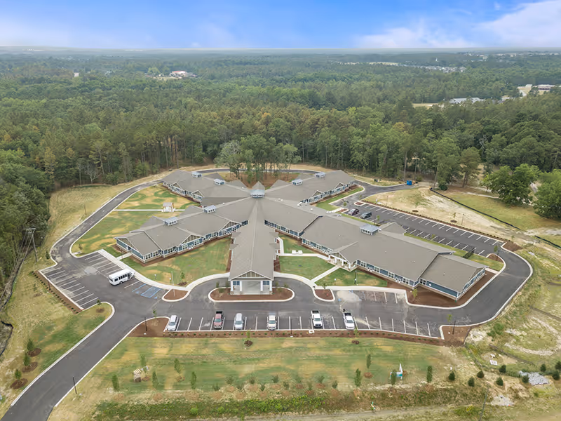 Aerial view of Lakeside Place Senior Living facility showing a large, single-story building with multiple wings arranged in a star-like pattern surrounded by parking lots and green wooded areas.