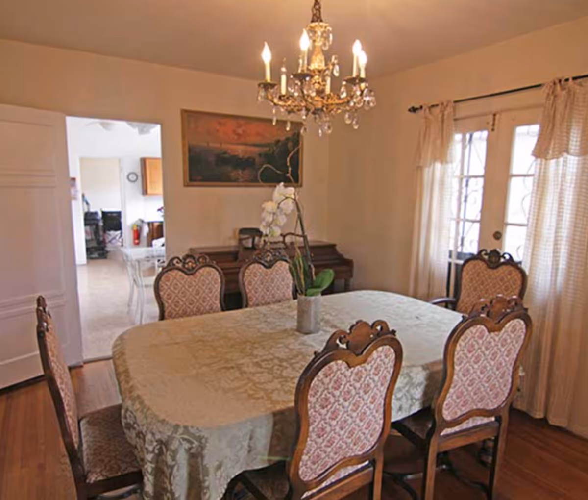 A dining room with a rectangular table covered by a patterned tablecloth and surrounded by six upholstered wooden chairs. A chandelier with lit bulbs hangs above the table. There is a potted orchid plant in the center of the table. The room has wooden flooring, light-colored walls, a painting on the wall, and a set of French doors with curtains.
