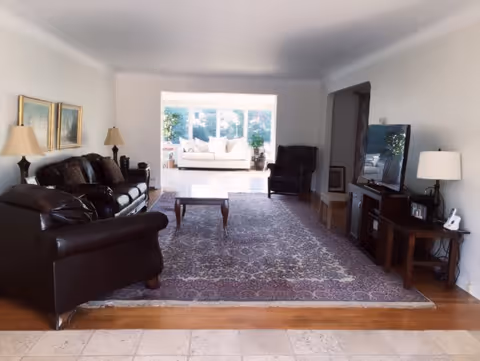 Spacious living room with dark leather furniture, wooden coffee table, area rug, and bright sunroom beyond.